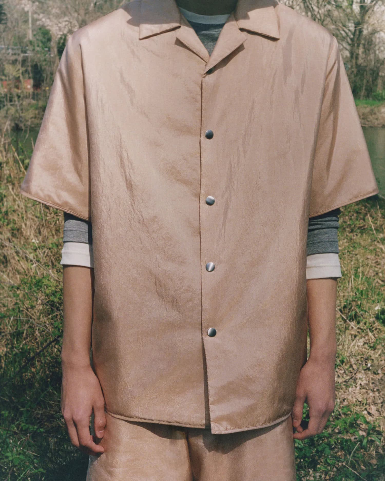Person in a beige short-sleeved button-down shirt with matching shorts standing outdoors, showcasing fashion and style in a natural setting.