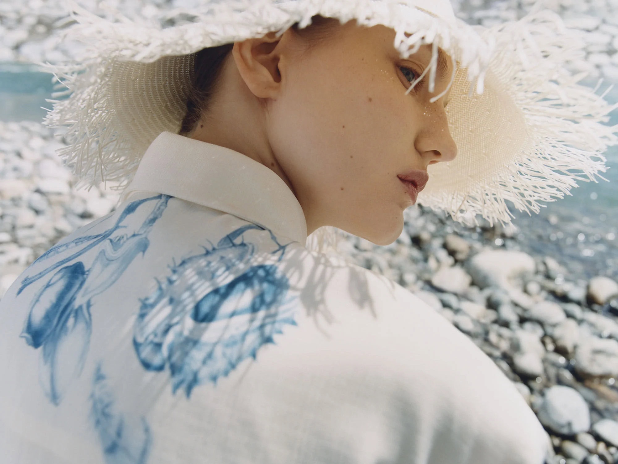 Side profile of an adult woman with blond hair wearing a white jacket with a blue floral pattern and a wide-brimmed hat in nature.