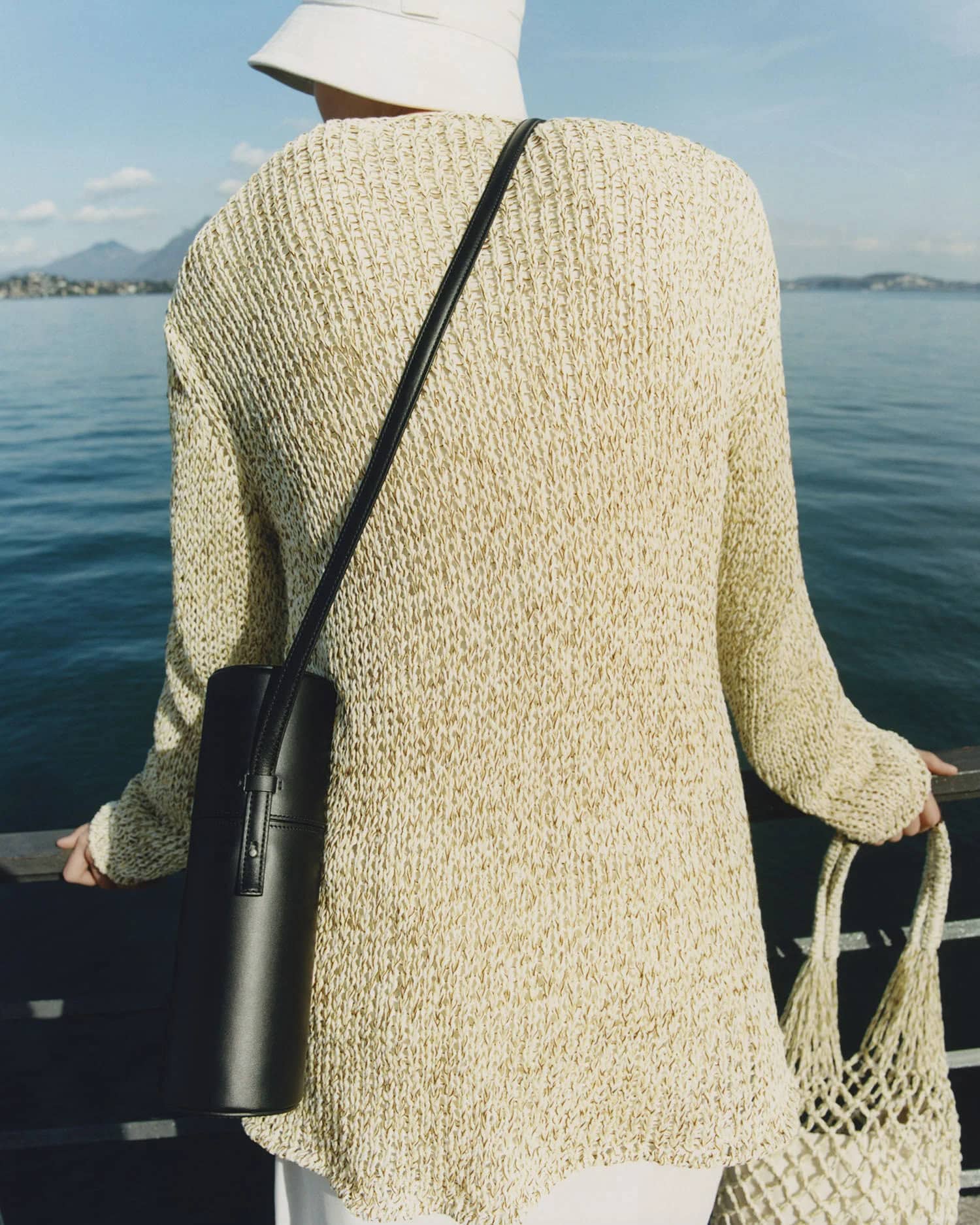 A person stands by a lake wearing a tan knit sweater, bucket hat, black purse, and holding a beige net bag, with mountains in the distance.