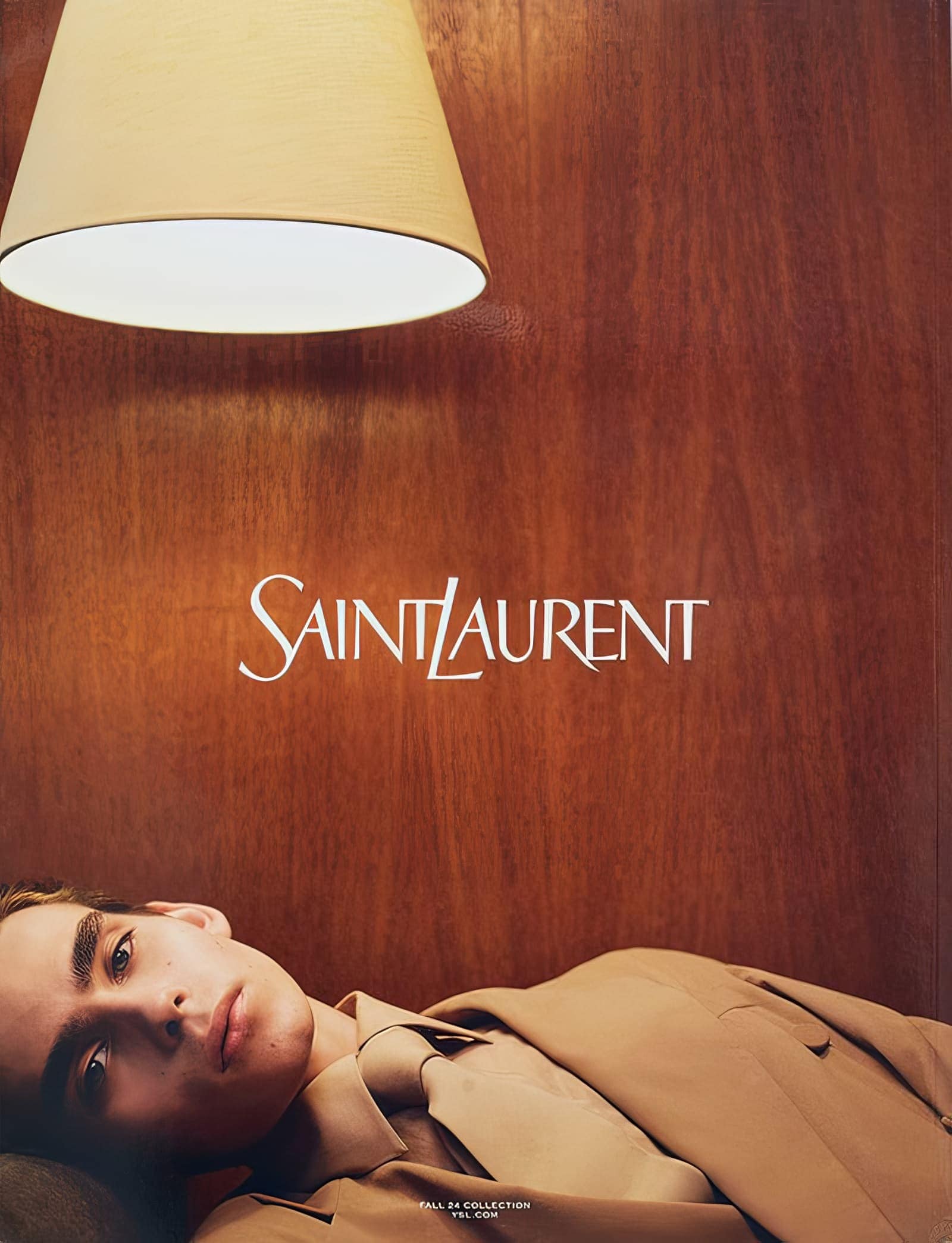 Fashion editorial featuring a young adult male model wearing a tan suit, lying under a lamp in front of a wood panel backdrop.