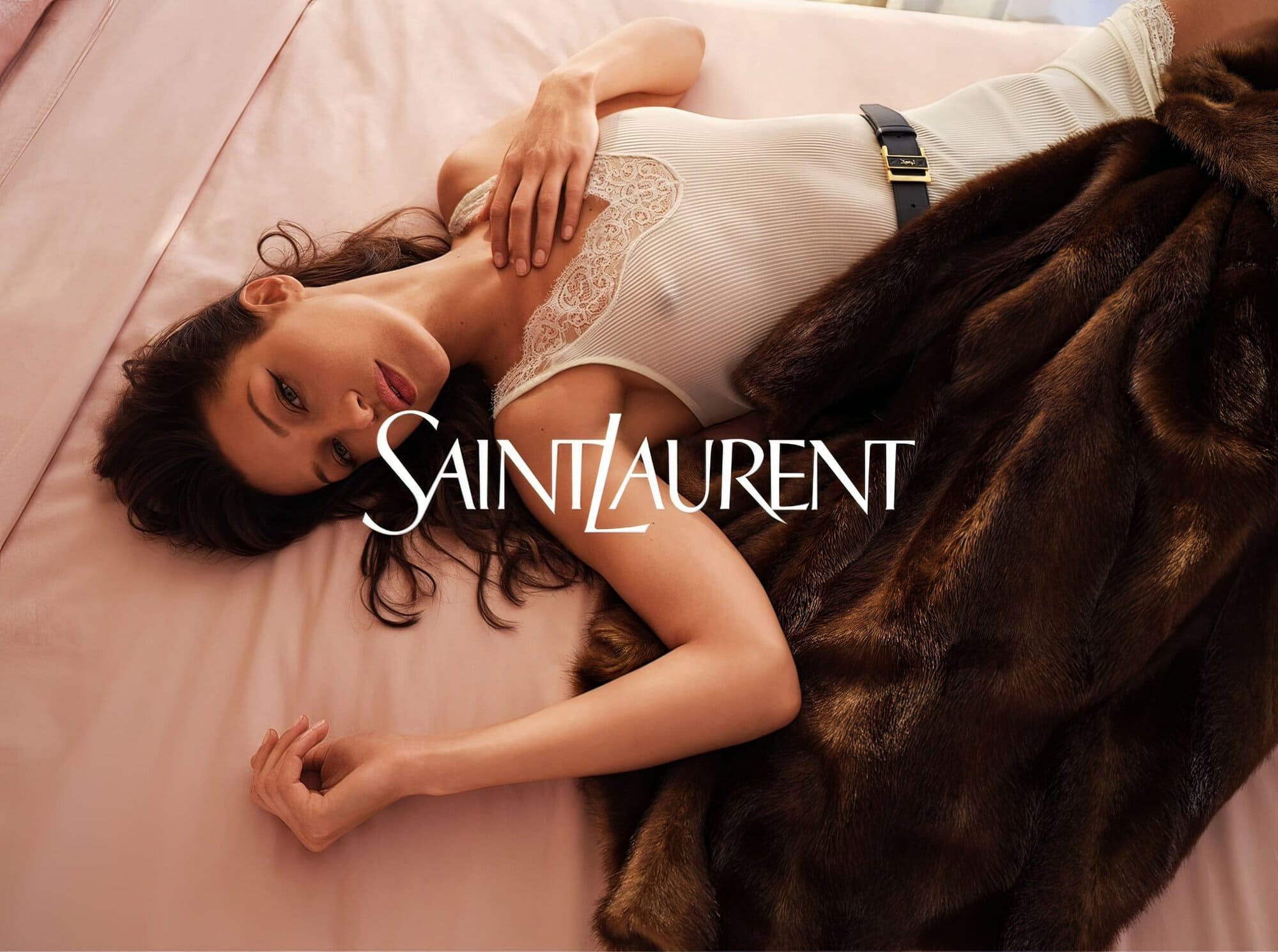 Brunette model in Saint Laurent ad, reclining on pink sheets with a fur coat, looking at the camera with a neutral facial expression.