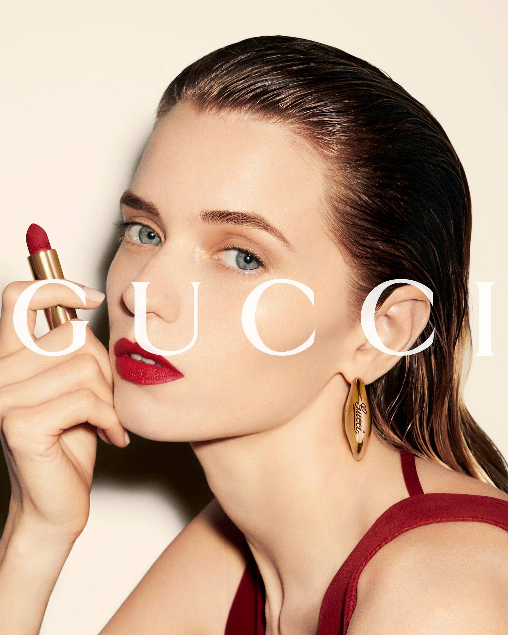 Beauty shot of a woman with red lipstick holding a tube of lipstick, wearing an earring and sleeveless top against a cream background.
