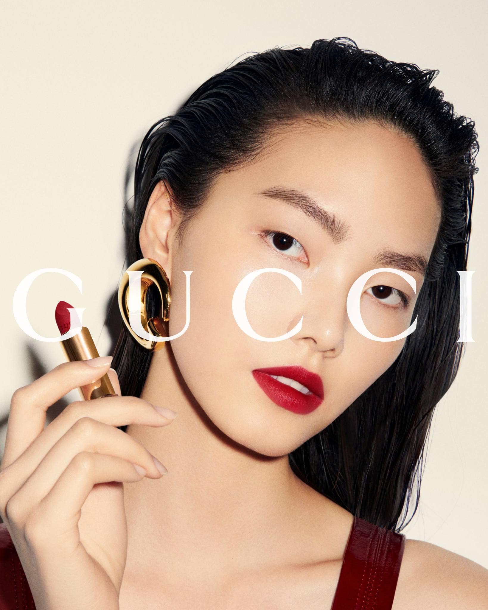 Close-up of an Asian female model in a beauty ad featuring Gucci lipstick, gold earrings, and slicked-back dark hair against an off-white backdrop.