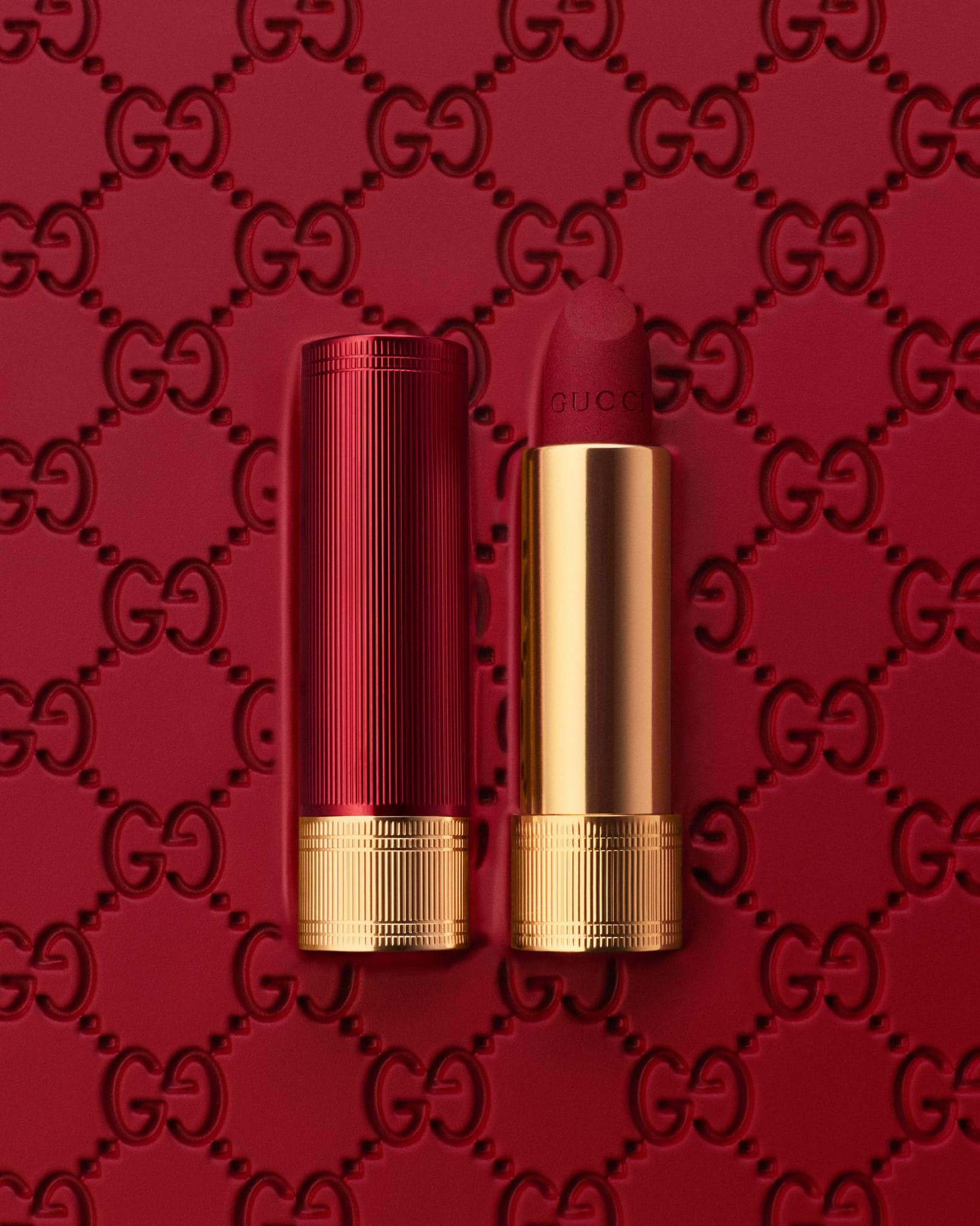 Close-up of a Gucci lipstick with its lid open, displayed on a red surface featuring the Gucci logo pattern for a beauty advertisement.