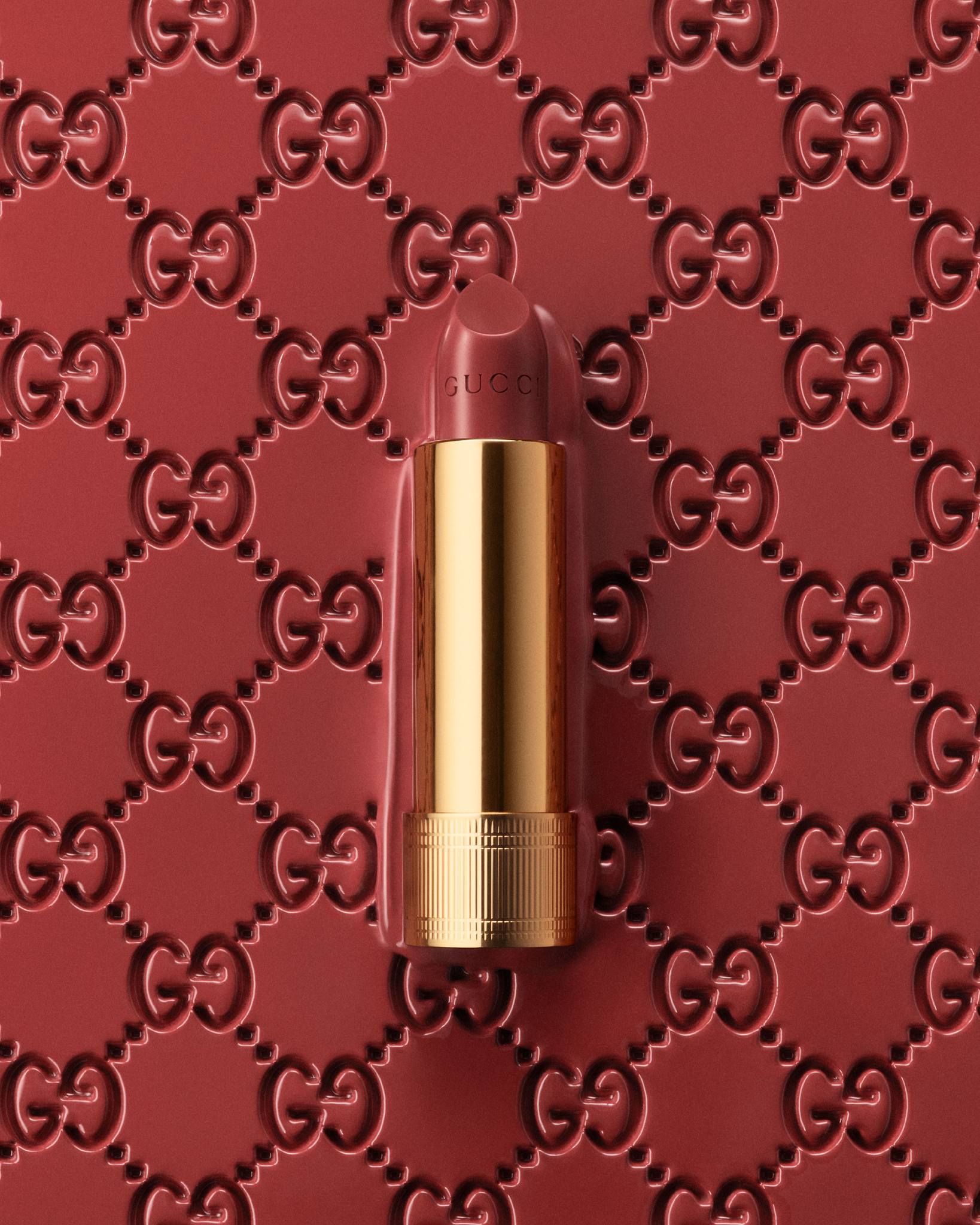 Close-up studio shot of a Gucci lipstick tube in a gold case against a rose-colored background with a repeated Gucci logo pattern.