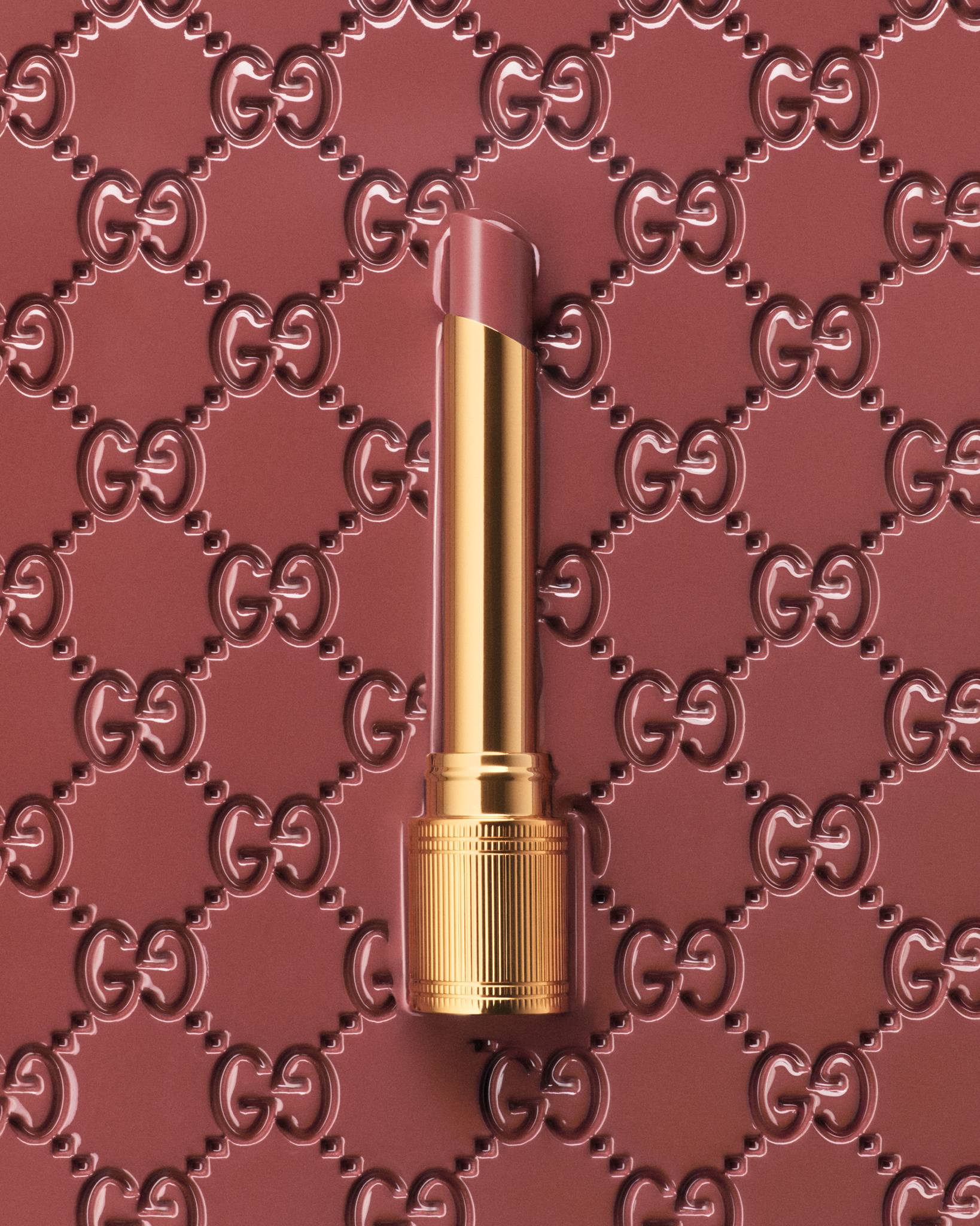 Gucci lipstick with gold tube and mauve color against background with repeating embossed Gucci logo pattern, top-down beauty product shot.