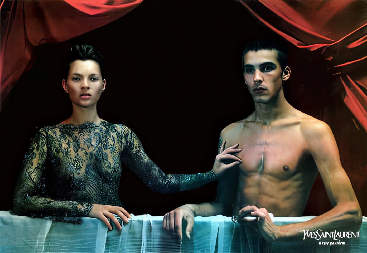 Fashion shot with a fair-skinned male model and fair-skinned female model in dark lacy top, set against a dark background and red curtains.