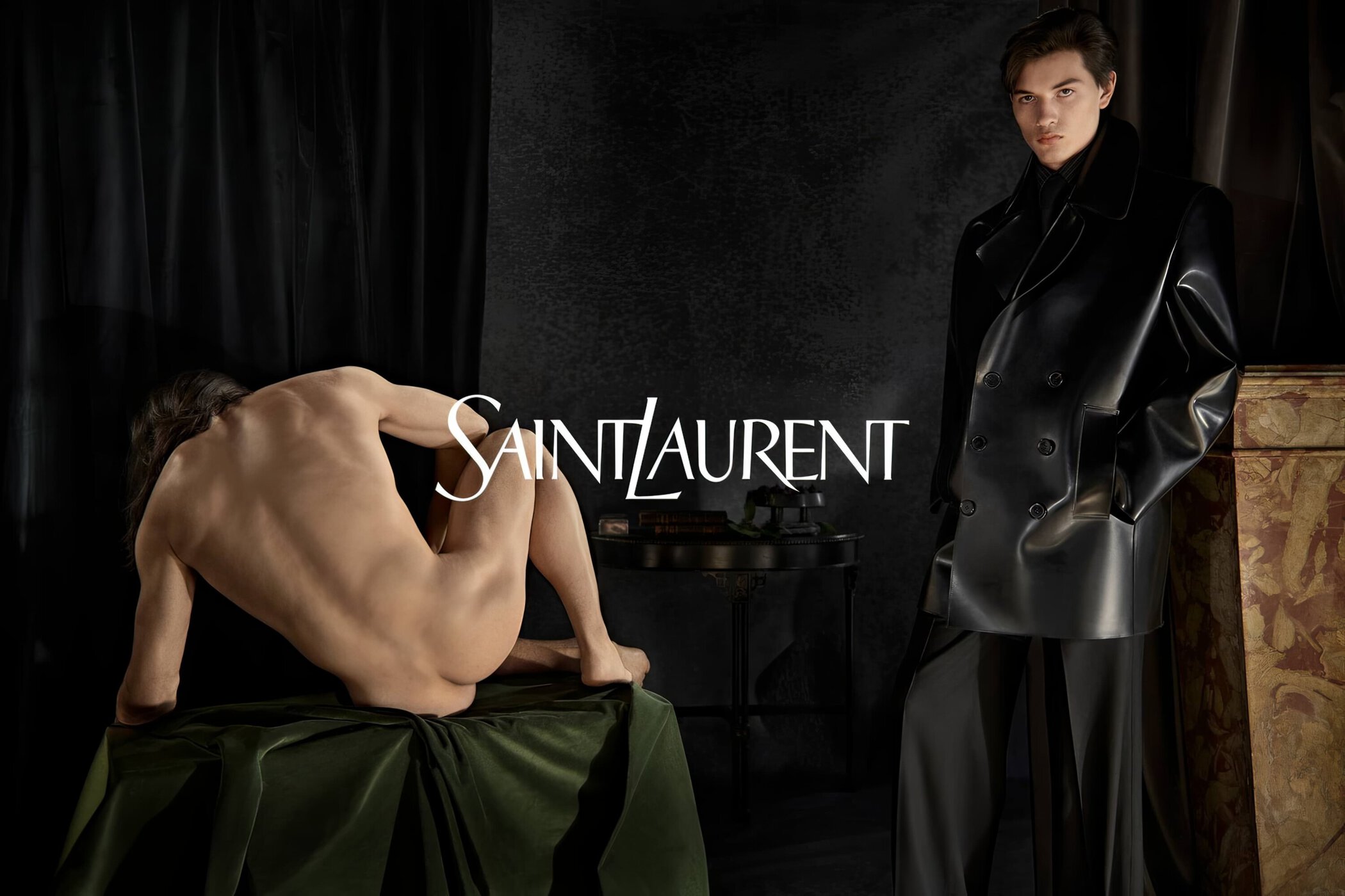 Two male models pose in a studio setting, one nude on a velvet cloth, the other in a black leather coat and trousers for Saint Laurent.