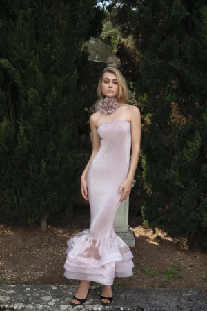Elegant woman in pink strapless gown with a floral choker posing in a garden with green foliage and stone architecture in the background.