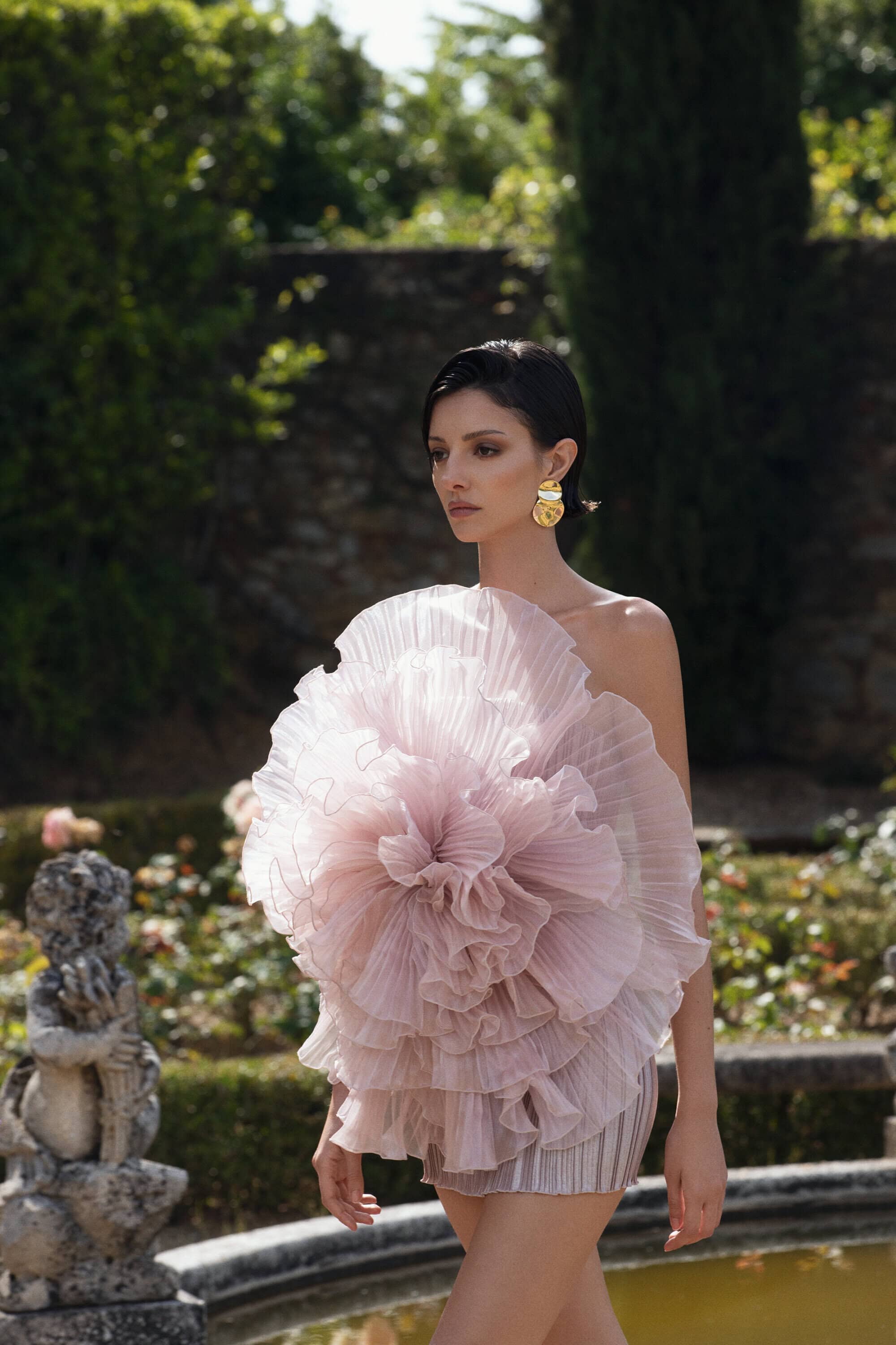 Woman wearing a large ruffled pink pleated dress standing outdoors in a garden for Lidee Woman