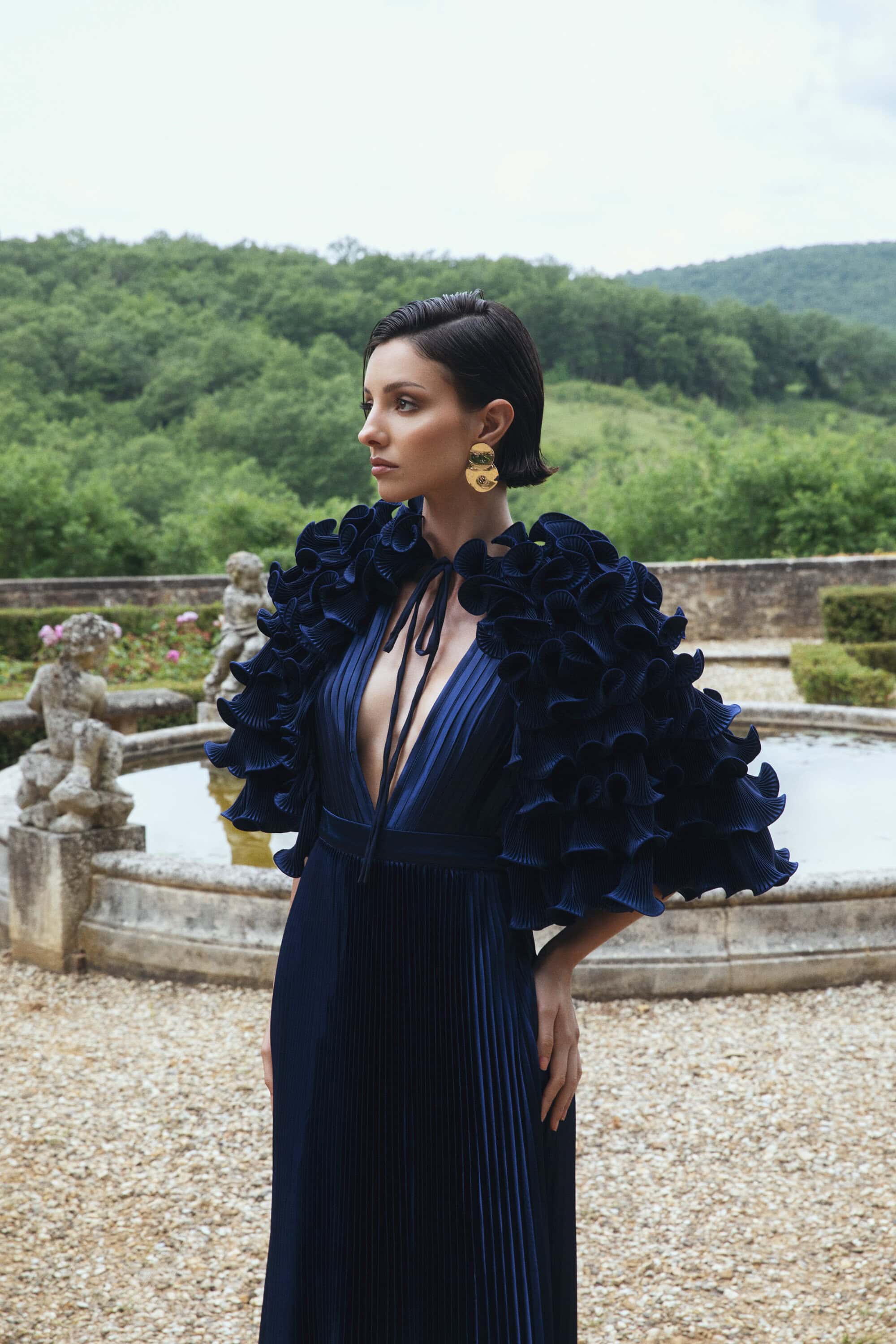 An elegant woman in a ruffled, navy blue gown poses in a garden with a fountain, trees, and sky in the background, capturing a serene outdoor moment.