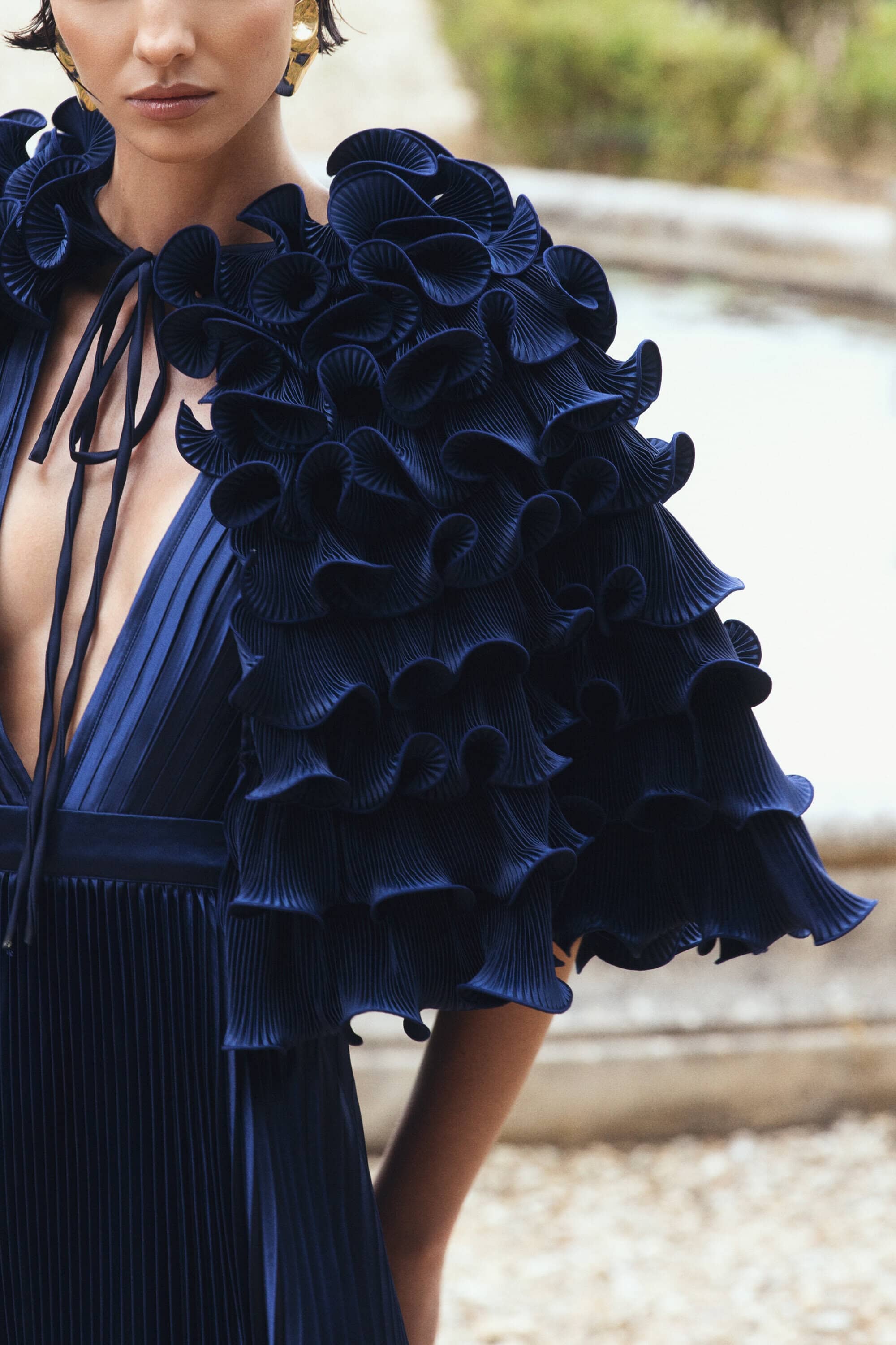 Close-up of a fair-skinned adult female model wearing a dark blue pleated dress with ruffle details, posing outdoors with natural lighting.