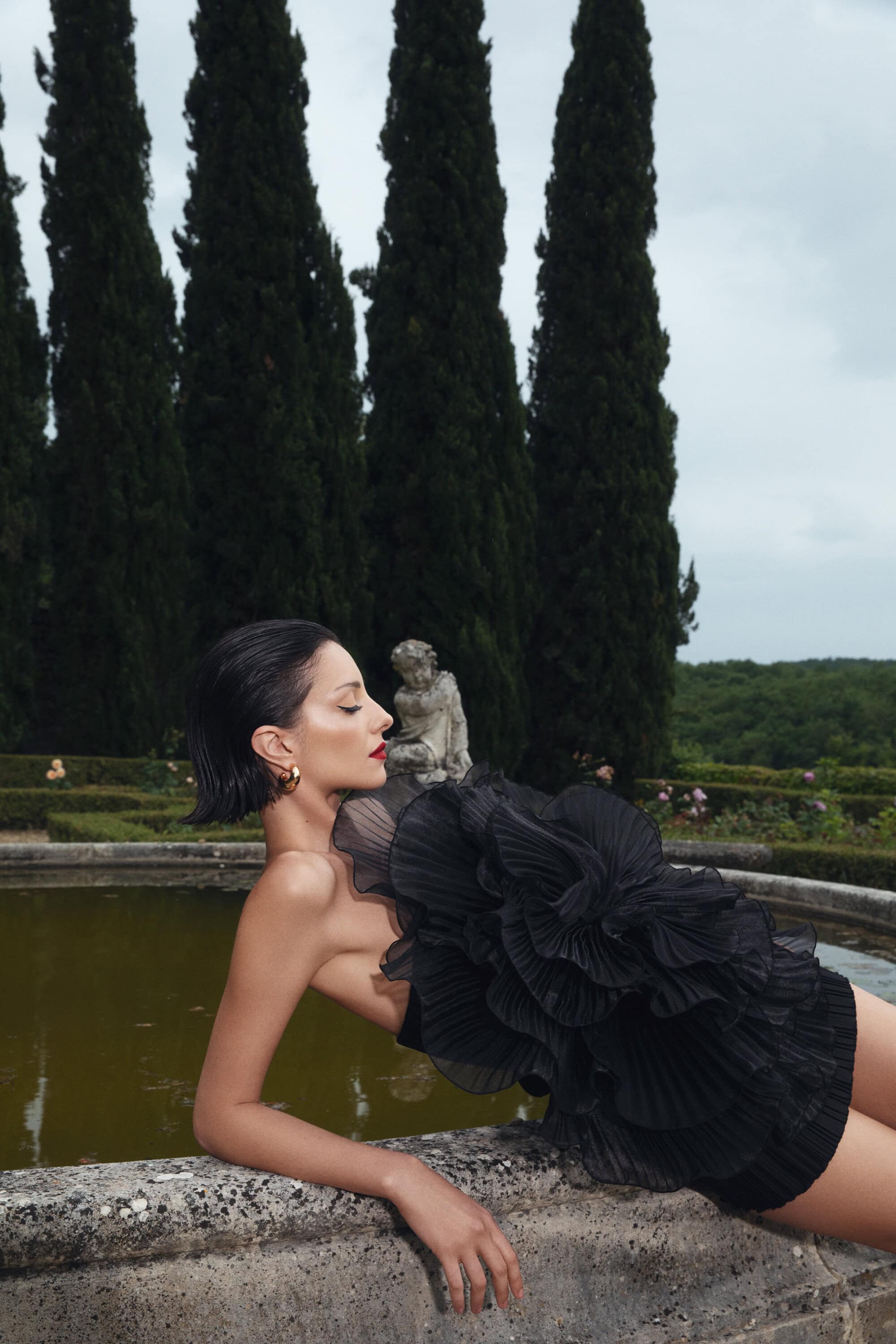 Adult woman lies on a stone wall in a black ruffled dress near a pond and formal garden, framed by tall dark green trees against a cloudy sky.