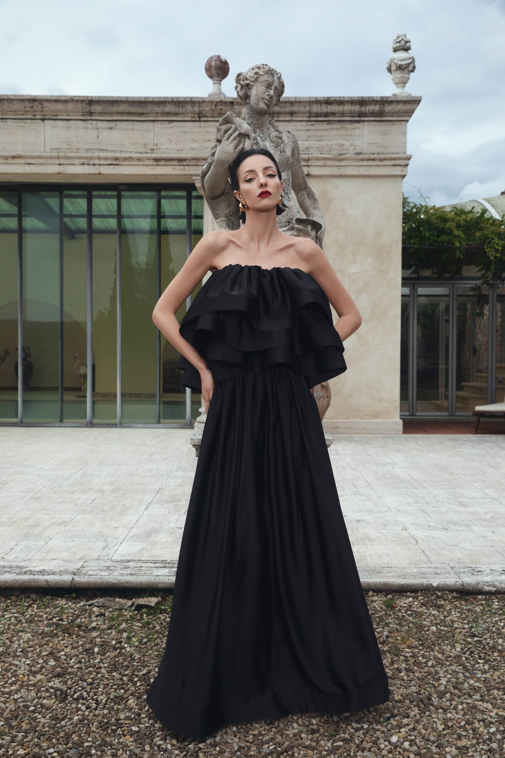 A woman poses in a black dress with a strapless ruffle bodice, standing in front of a sculpture in an outdoor garden setting.