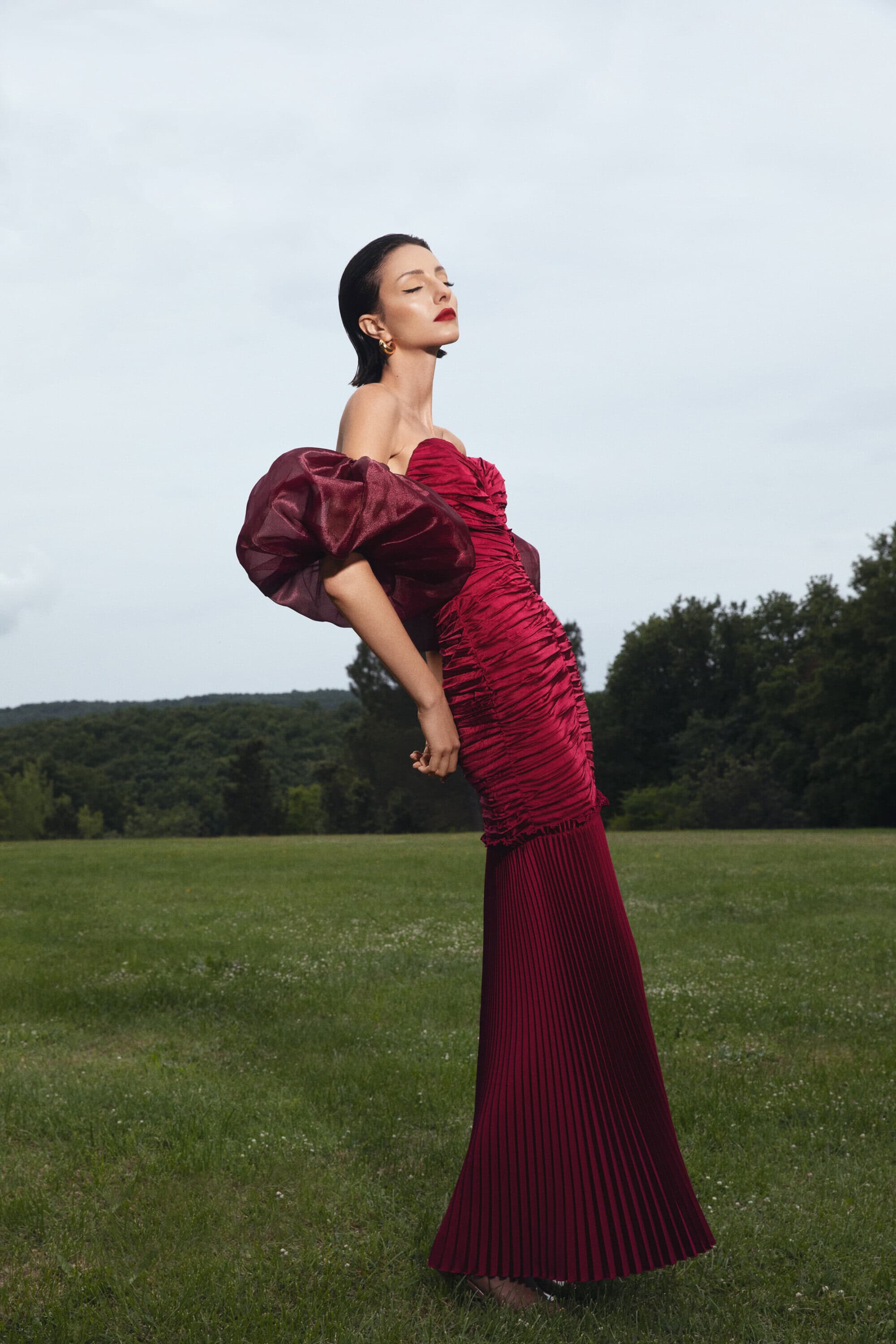 Woman in a red gown poses in a grassy field with trees. The dress has a sweetheart neckline and puffy sleeves against a cloudy sky.