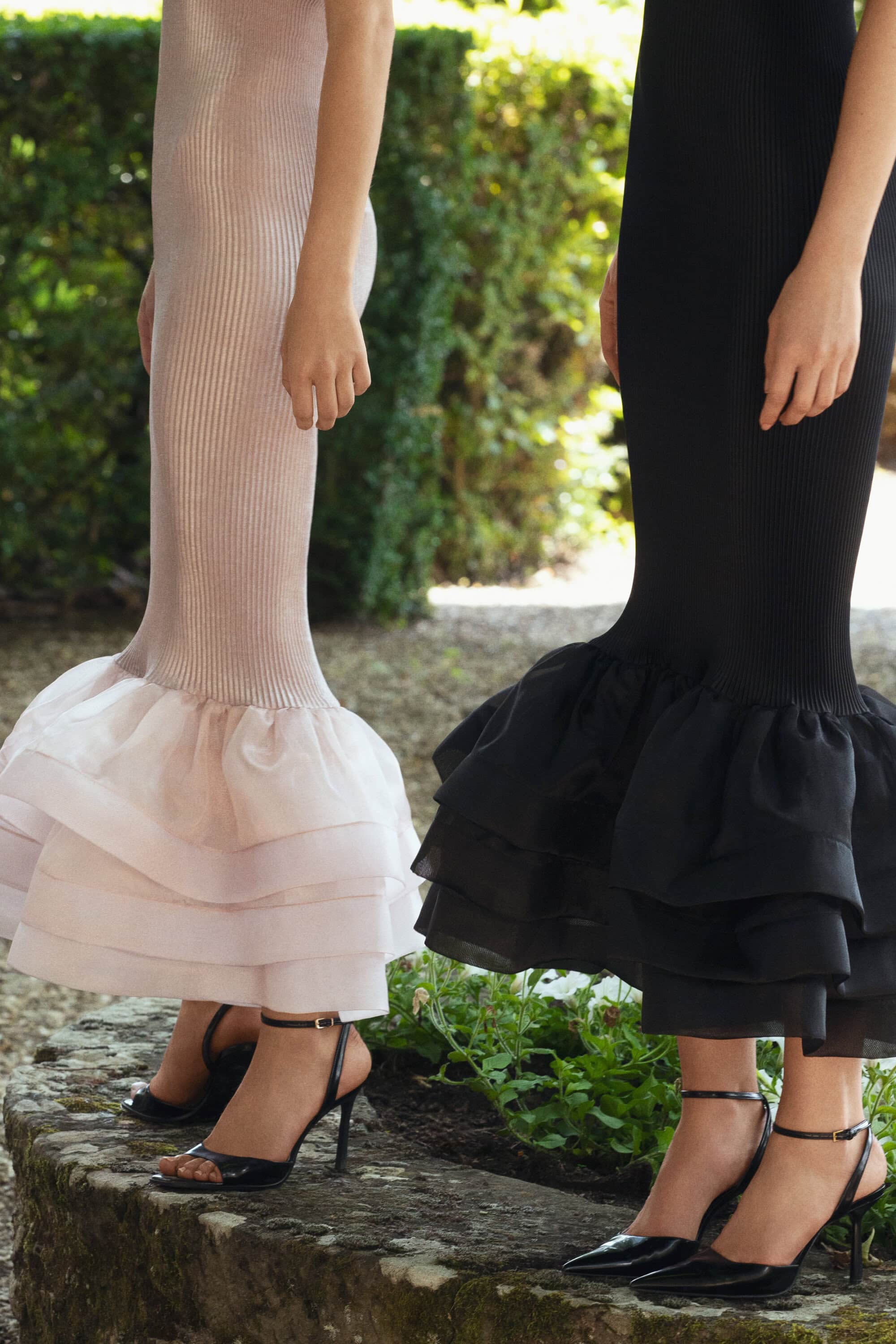 Two women model elegant ribbed dresses with layered tulle ruffles and ankle-strap heels, posing outdoors against a green hedge backdrop.