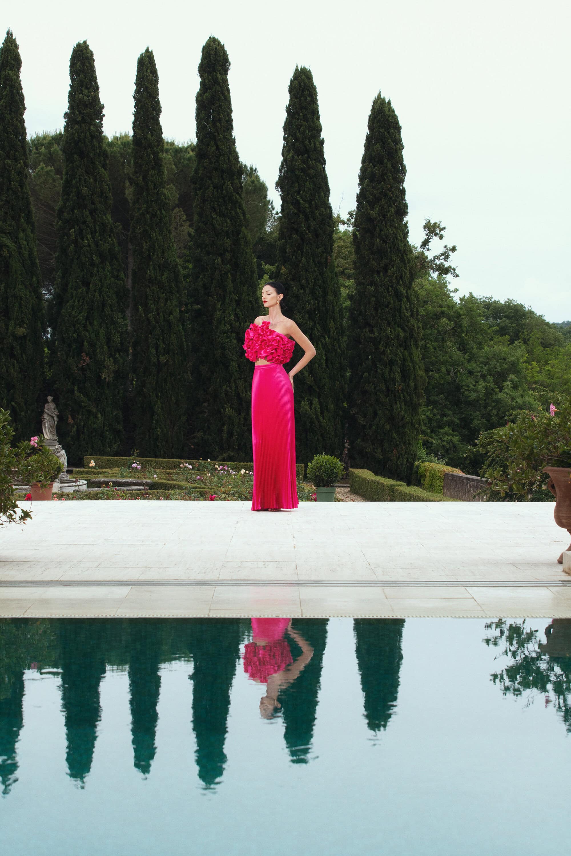 A woman in a pink dress stands poolside, reflected in the water with green trees in the background under an overcast sky.