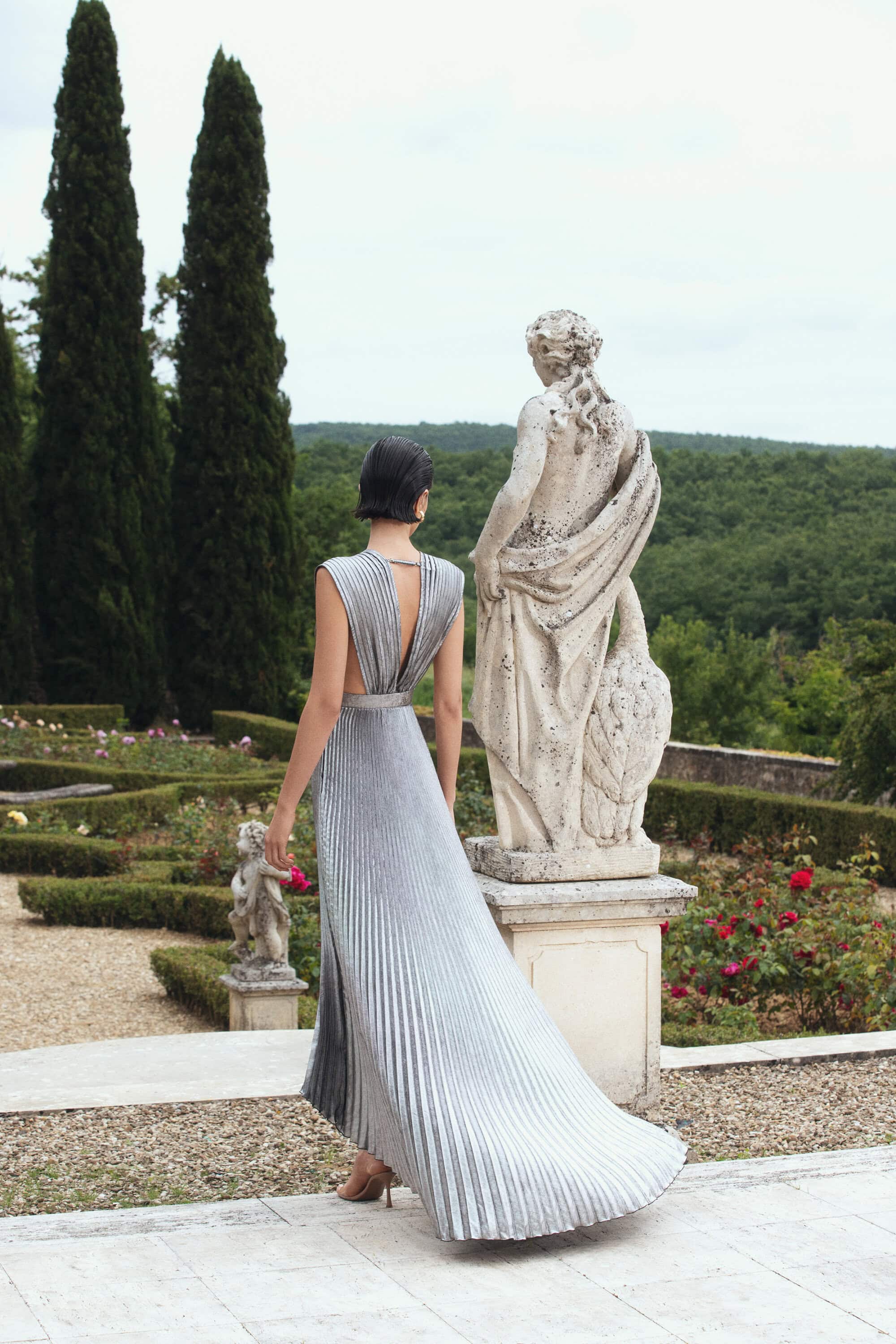 Back view of a stylish adult woman wearing a pleated gown in a garden setting with classical statues and greenery in the background.