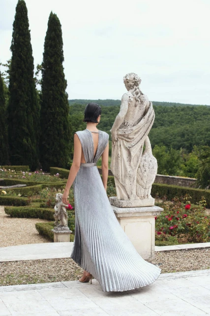 Back view of a stylish adult woman wearing a pleated gown in a garden setting with classical statues and greenery in the background.