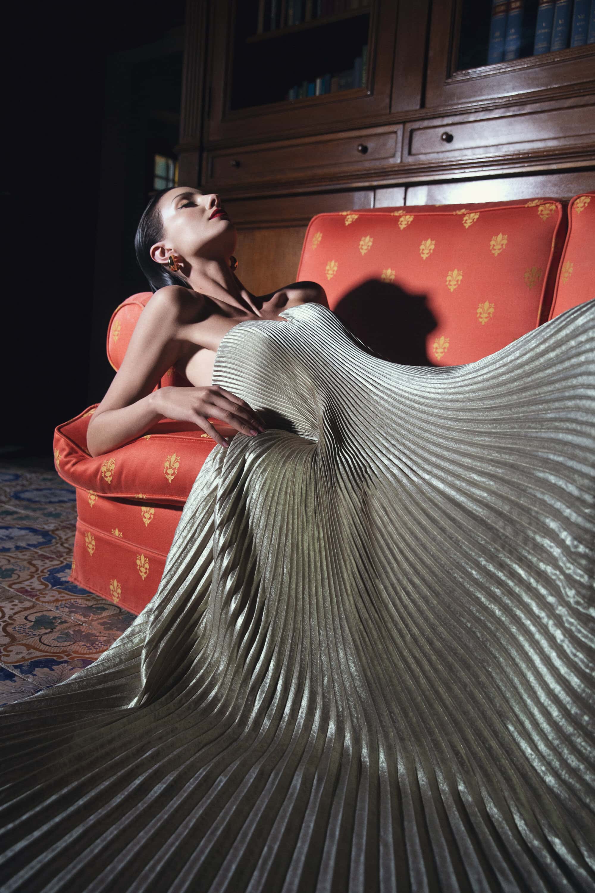 Woman in shimmering pleated gown reclining on an orange couch in a dimly lit room with dark wood bookcase behind her.