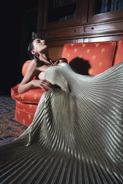 Woman in shimmering pleated gown reclining on an orange couch in a dimly lit room with dark wood bookcase behind her.