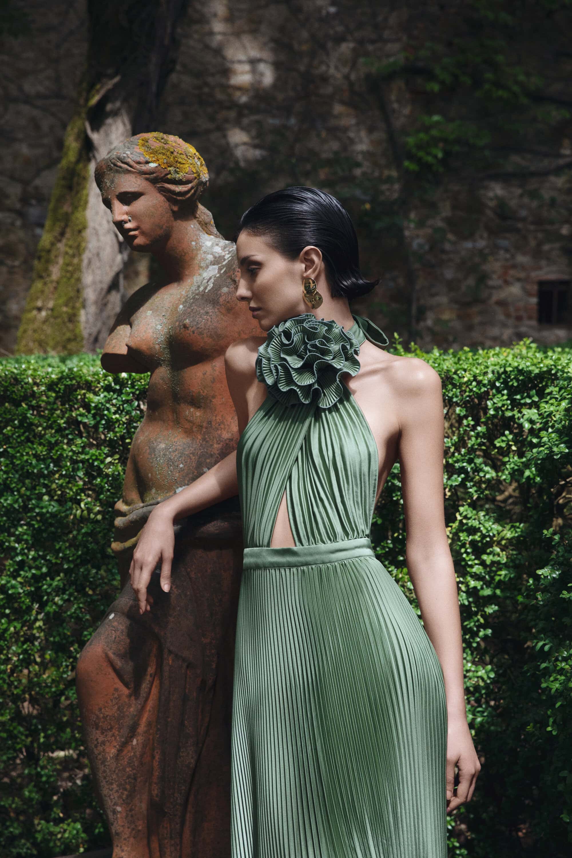 Woman in pleated green dress poses next to outdoor statue in lush garden setting, highlighting fashion and classical art fusion.