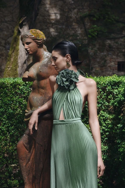 Woman in pleated green dress poses next to outdoor statue in lush garden setting, highlighting fashion and classical art fusion.