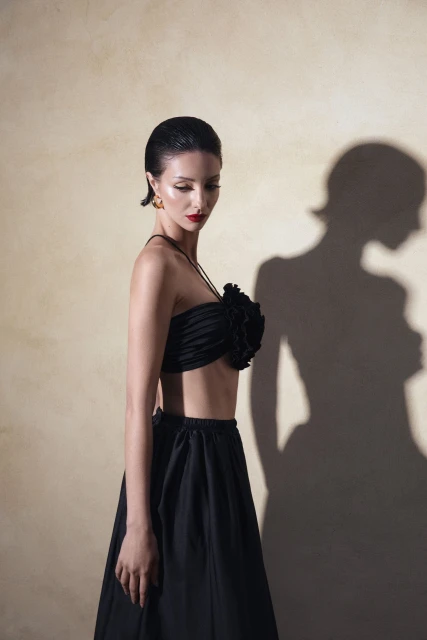 Adult female poses indoors in a black strapless crop top with a floral detail and a black skirt against a beige mottled background.