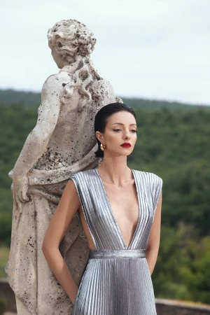 Adult woman in a silver dress poses by a stone statue outdoors in daytime, fashion photography. Elegant style with architectural backdrop.