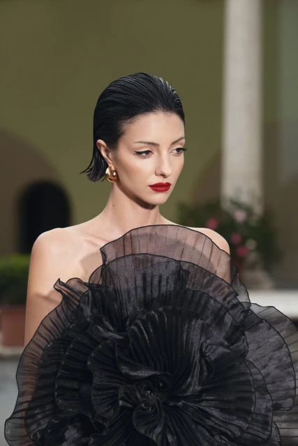Brunette woman poses in a black ruffled, floral dress with red lipstick, gold earrings, and slicked-back hair against an outdoor backdrop.