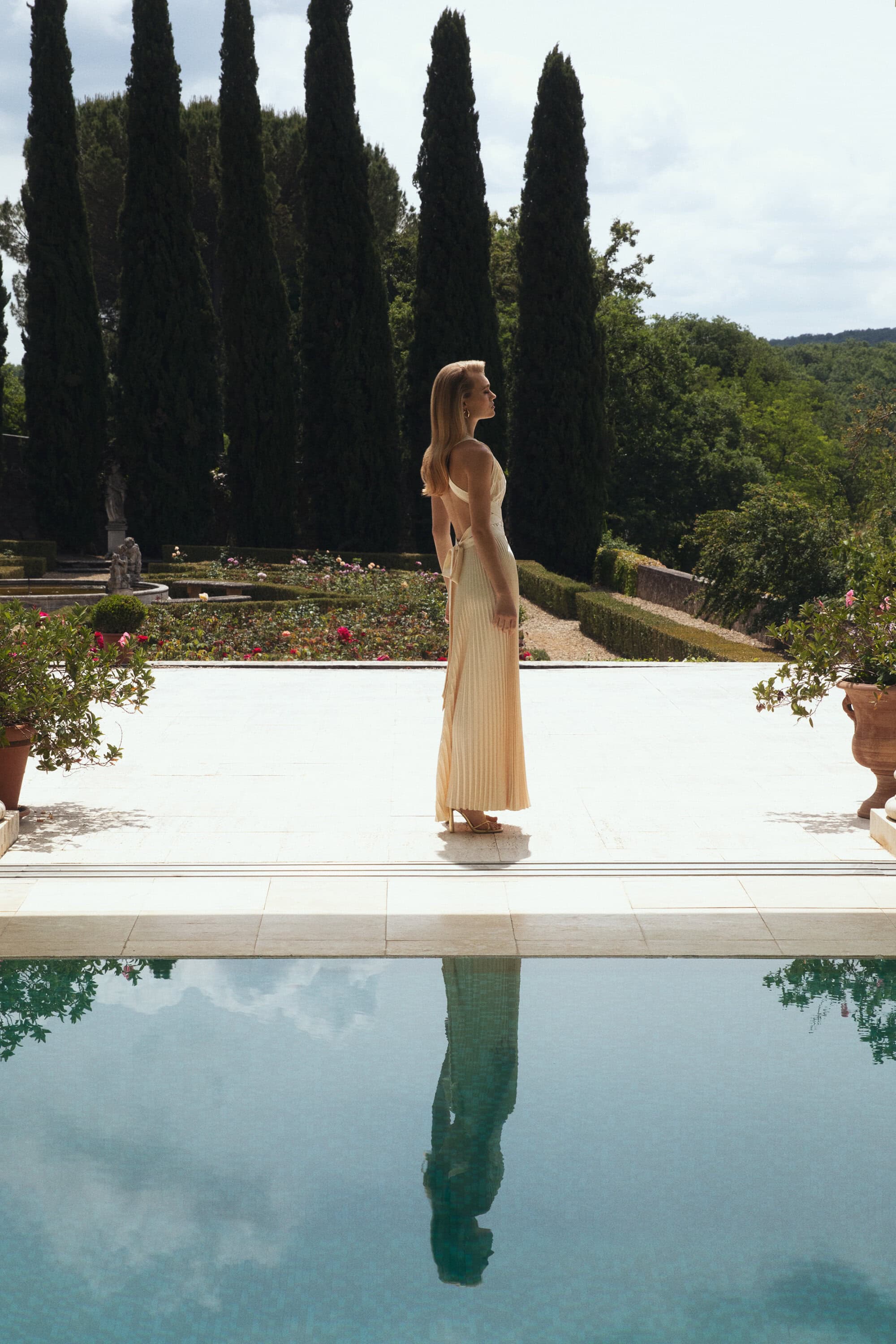 Woman in formal dress standing on a patio overlooking a reflecting pool and garden with cypress trees in the background.