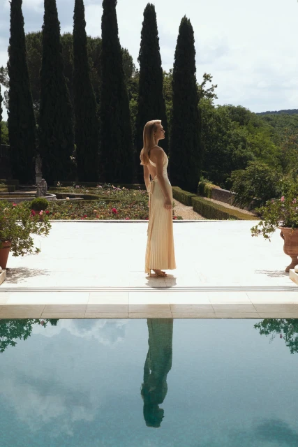 Woman in formal dress standing on a patio overlooking a reflecting pool and garden with cypress trees in the background.