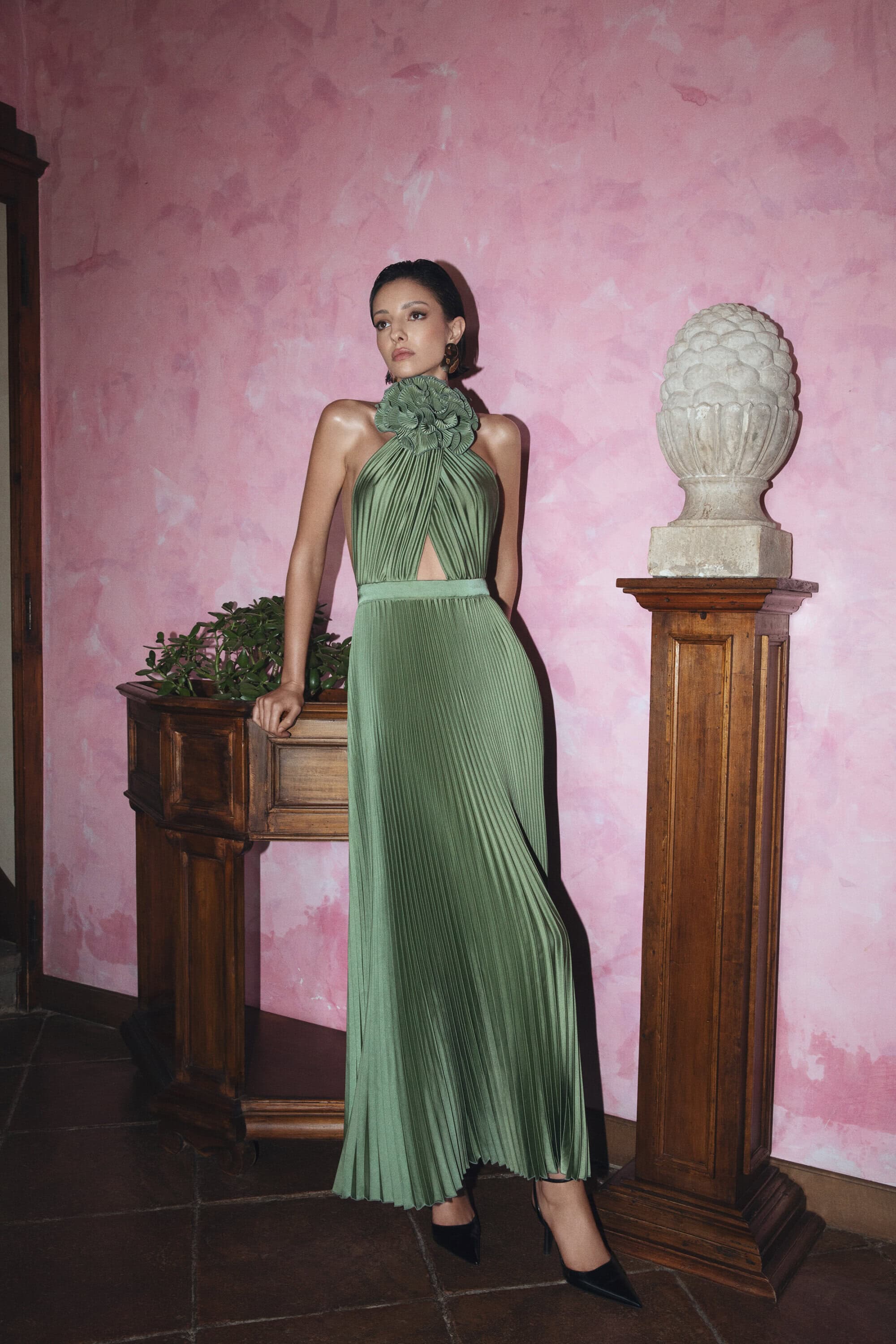 Elegant woman in a green pleated halter gown poses in front of a vintage wall, showcasing high fashion and classic beauty indoors.