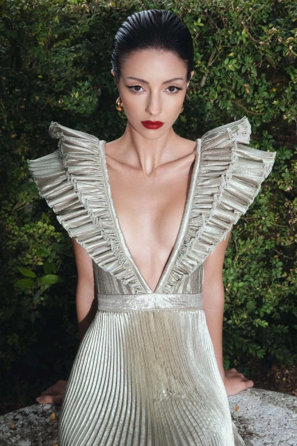 Elegant woman in a gold, floor-length gown with ruffles poses outdoors against a green leafy background for a fashion editorial shoot.