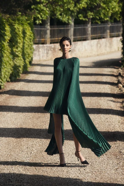 Woman wearing a draped, pleated emerald green gown and high heels stands on a path lined with trees and greenery in an outdoor setting.