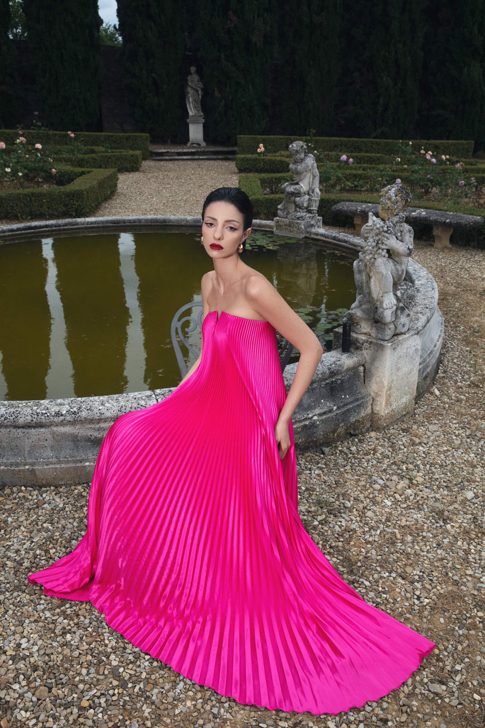 Woman poses in a vibrant pink strapless gown beside a water feature in a formal garden with manicured hedges and gravel pathways.