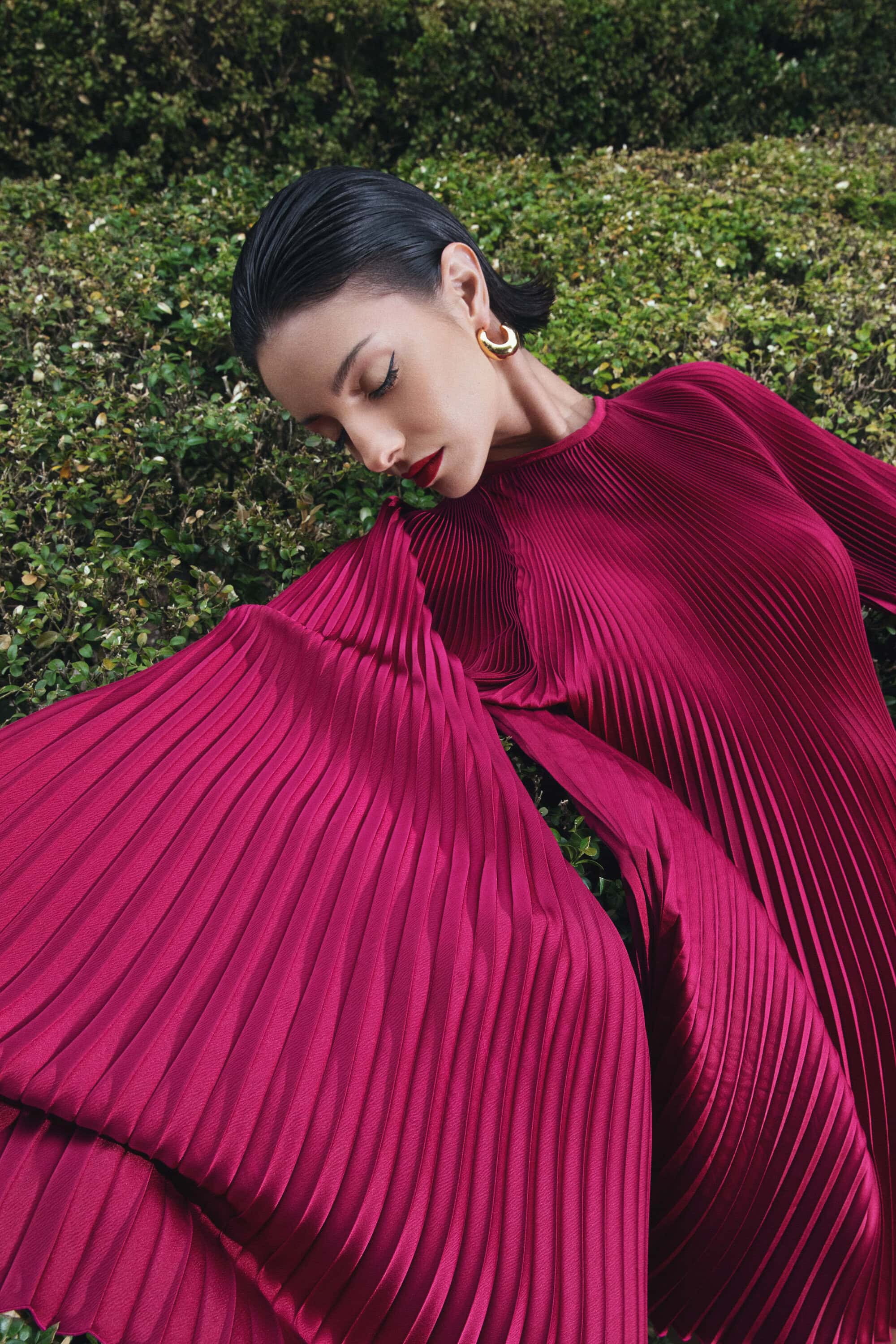 Elegant woman in flowing magenta pleated dress posing in lush green garden setting, accessorized with gold hoop earrings, head tilted down.
