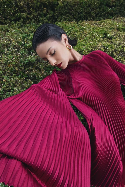 Elegant woman in flowing magenta pleated dress posing in lush green garden setting, accessorized with gold hoop earrings, head tilted down.