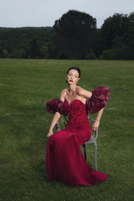 A woman in a red off-the-shoulder gown sitting on a chair in a green field in front of a forest under a cloudy sky.