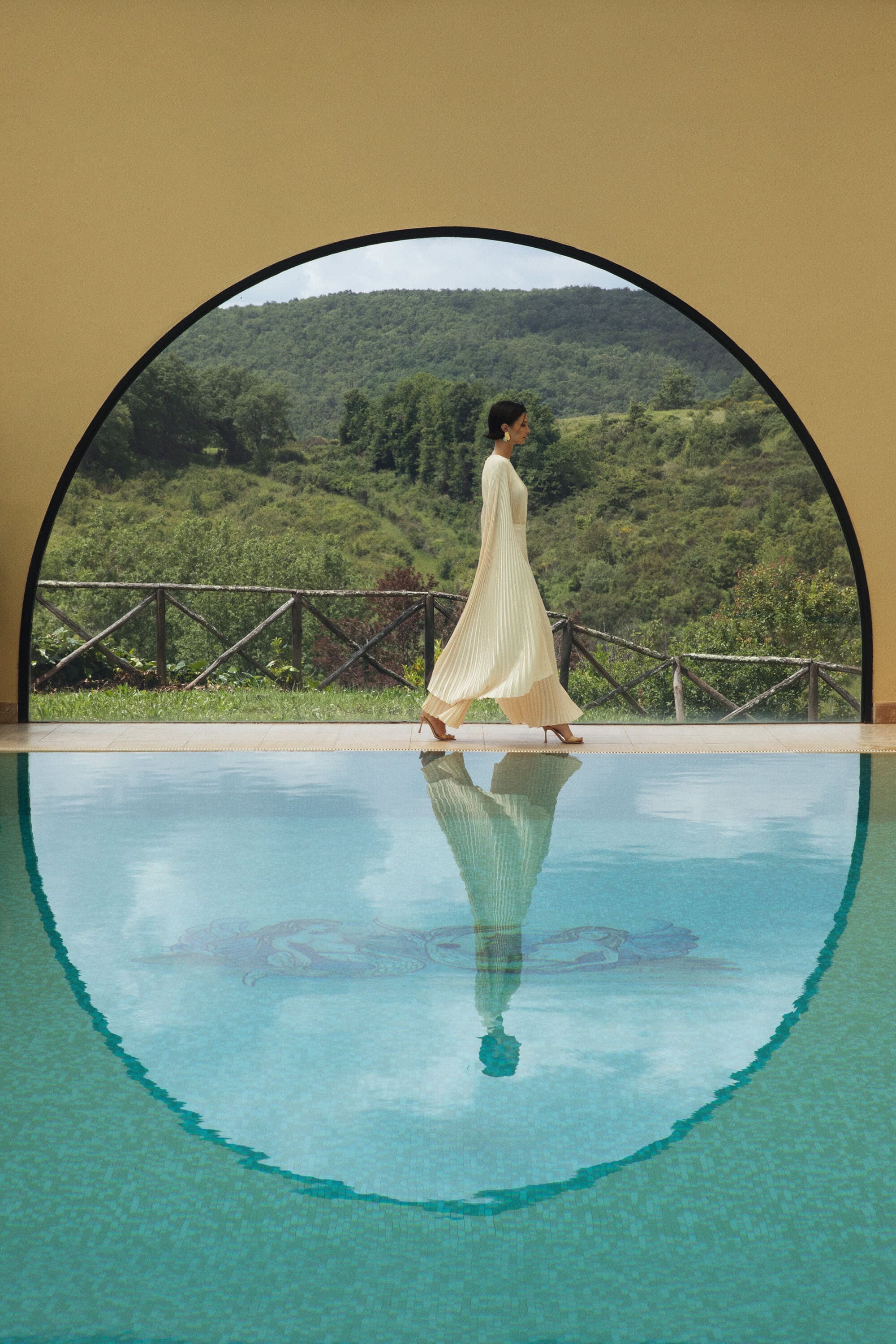 Adult woman in a cream-colored dress walks along a pool edge by a building with green hills visible through an arched opening.