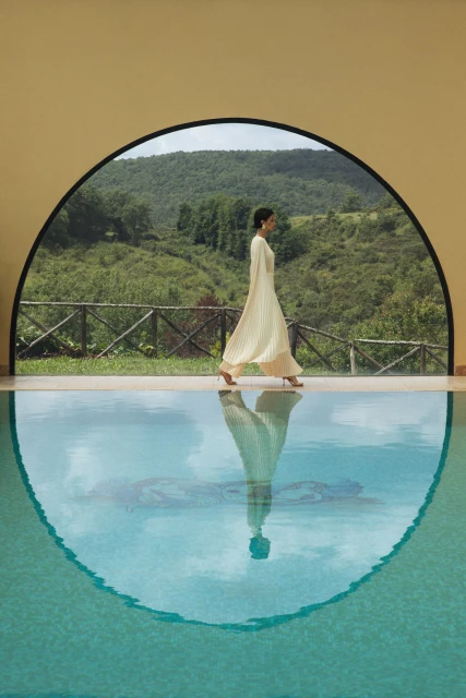 Adult woman in a cream-colored dress walks along a pool edge by a building with green hills visible through an arched opening.