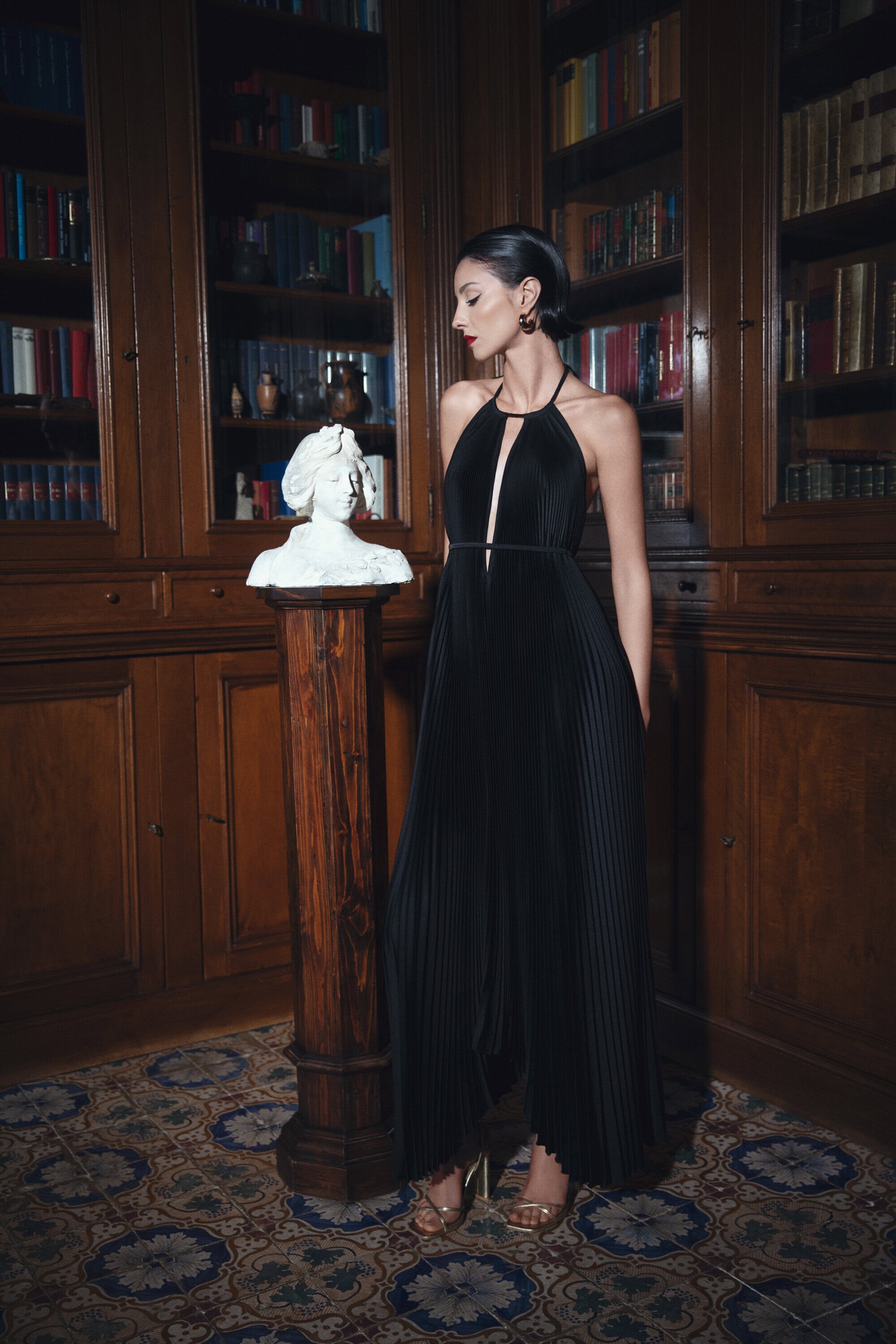 A woman in a black evening gown stands next to a bust in a library setting with classic architecture and tiled flooring.
