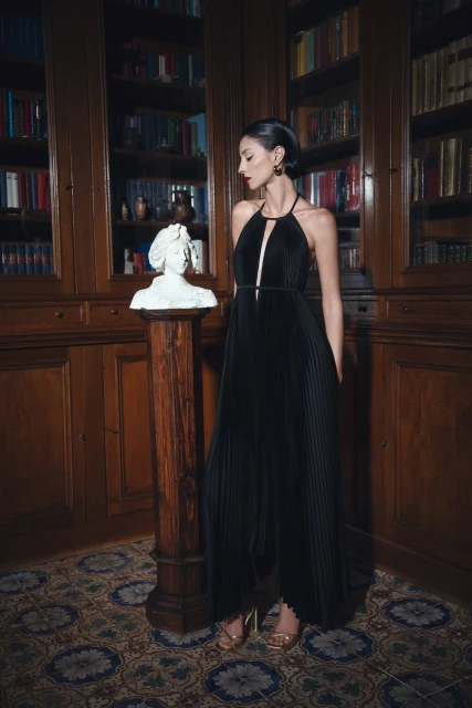 A woman in a black evening gown stands next to a bust in a library setting with classic architecture and tiled flooring.