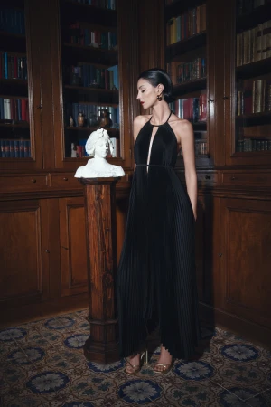 A woman in a black evening gown stands next to a bust in a library setting with classic architecture and tiled flooring.