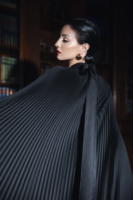 Elegant woman in flowing black dress with pleated texture, posed in profile view against a dark backdrop with a vintage bookcase.