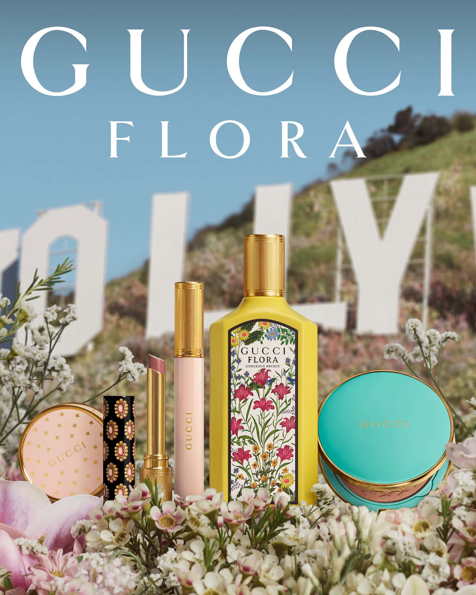 Gucci Flora perfume bottle, makeup compacts, and lipsticks arranged with flowers, set against the Hollywood sign under a bright blue sky.
