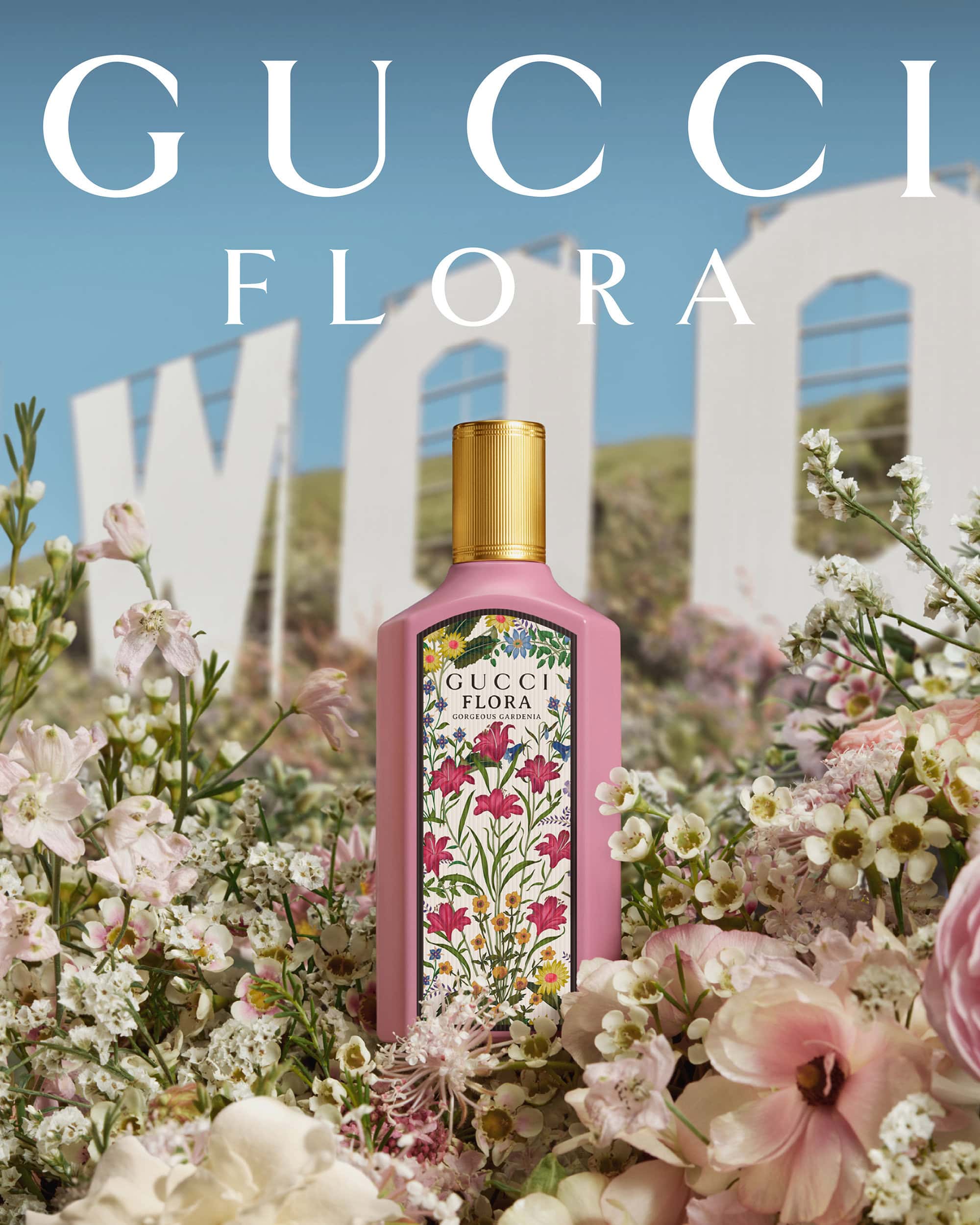 Gucci Flora perfume bottle stands surrounded by pink and white flowers against a backdrop of the Hollywood sign under a blue sky.