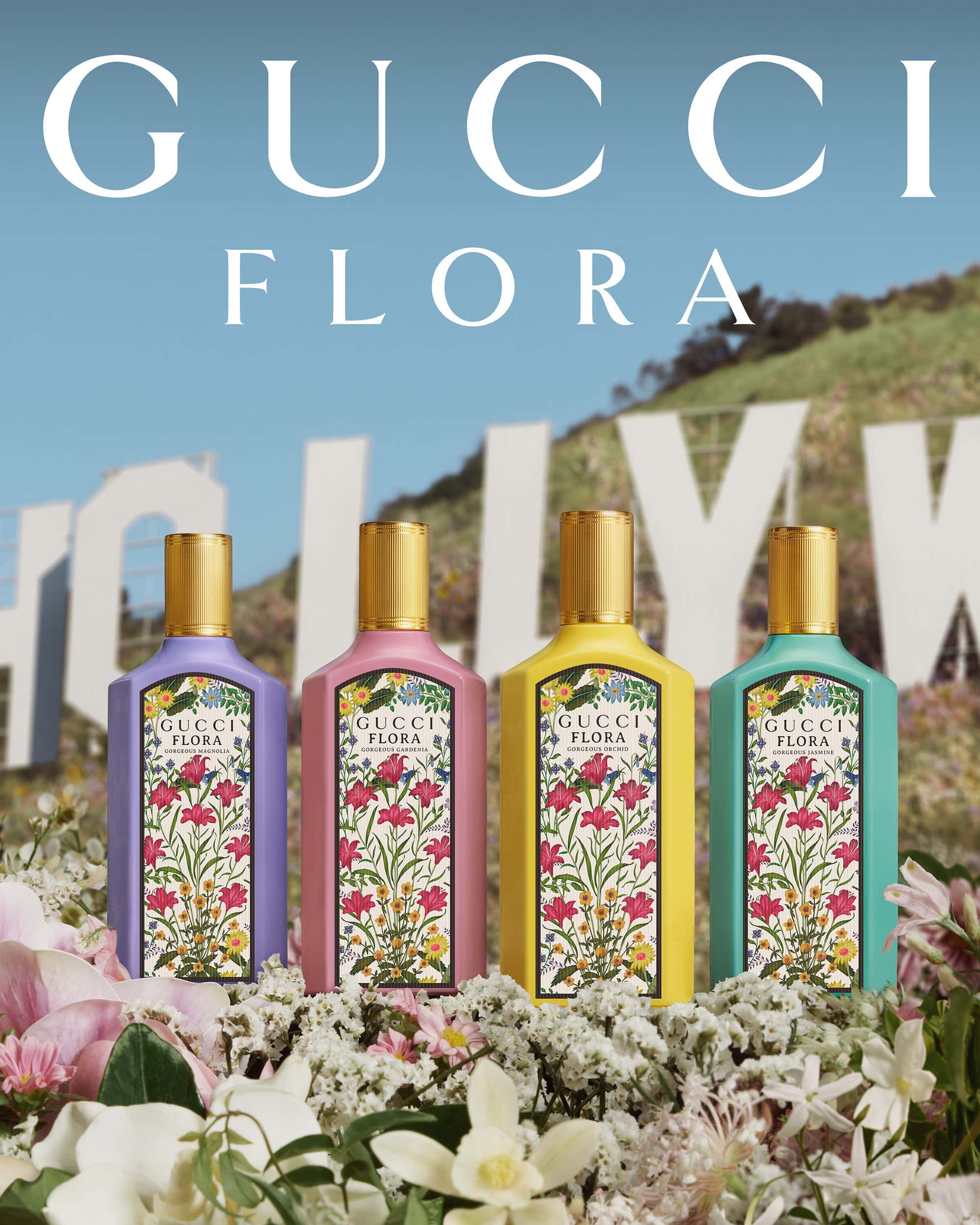 Four colorful Gucci Flora perfume bottles are arranged among white flowers with the Hollywood sign in the background on a sunny day.