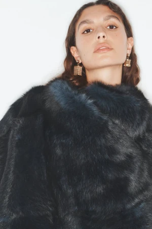 Brunette woman posing in a studio wearing a black fur coat and gold cube-shaped earrings, looking directly at the camera.