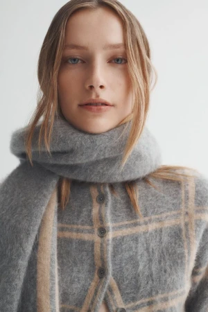 Close-up of a woman with blue eyes wearing a grey scarf and patterned sweater, looking directly at the camera in a studio setting.